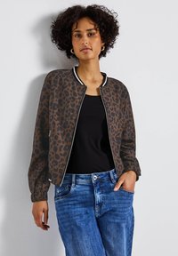 Hnědá bomber bunda s leopardím potiskem, s černým a bílým žebrovaným límcem, zapínáním na zip a nařasenými rukávy, v kombinaci s modrými džíny.