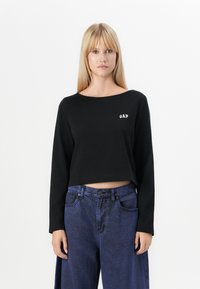 Svart kortärmad långärmad topp med rund halsringning och en vit "GAP"-logotyp på vänster bröst. Bärs med hög midja blå denimjeans.