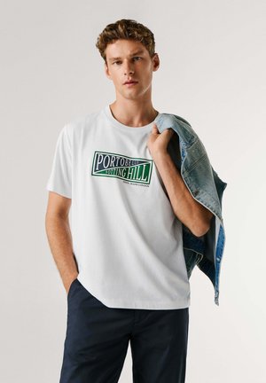 Maglietta bianca in cotone con testo verde "PORTOBELLO NOTTING HILL." Il modello tiene una giacca di jeans chiara appoggiata su una spalla. Pantaloni scuri.