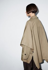 Übergroße khakifarbene Jacke aus leichtem Material, mit hohem Kragen, Knopfakzenten an den Ärmeln und einer lockeren, drapierten Silhouette.