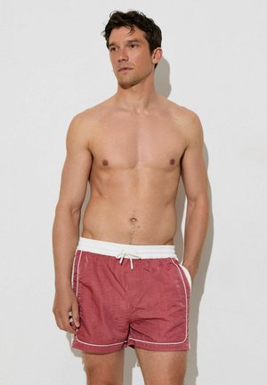 Man zonder shirt met kort donker haar, die rode zwemshorts met witte rand en tailleband draagt, staand tegen een effen lichte achtergrond.