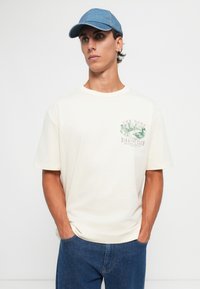 Ung mann iført en kremfarget T-skjorte med grønt fuglegrafikk og teksten "The Gant Birding Club," blå jeans og en lys blå baseballcaps, med hendene i lommene.