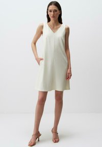 Abito senza maniche di un beige chiaro con scollatura a V, caratterizzato da una vestibilità morbida, due tasche laterali e una texture liscia.