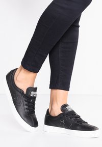 Personne portant des baskets en cuir Converse noires avec des semelles blanches et un pantalon skinny noir sur un fond blanc uni.