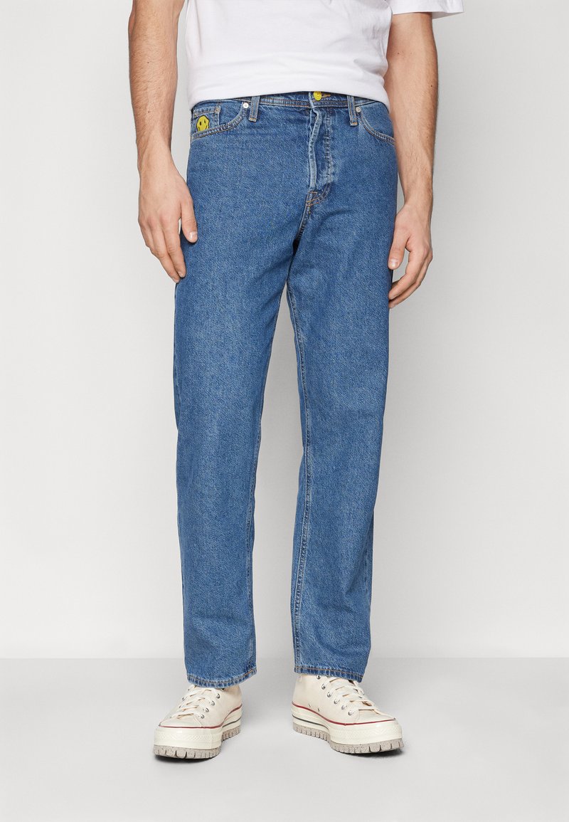 Blue denim jeans with a relaxed fit, featuring yellow hardware accents and visible stitching. Paired with white sneakers.
