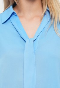 Ανοιχτό μπλε μπλούζα με γιακά και δεμένο λεπτομέρεια στον λαιμό. Το υλικό έχει μαλακή, ομαλή υφή με ελαφριά λάμψη.