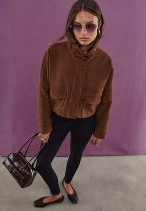 Veste en suédine marron avec un col montant et des poches avant, associée à des leggings noirs et des ballerines marron foncé, tenant un petit sac à main.