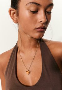 Jeune femme portant un top halter marron et un collier en chaîne dorée avec un pendentif en ambre carré, regardant vers le bas et vers sa droite.