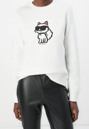 Osoba ubrana w białą bluzę z nadrukiem kreskówkowego kota w czarnych okularach przeciwsłonecznych na piersi oraz czarne spodnie z faux skóry.