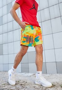 Colorful mesh shorts featuring a tie-dye pattern in orange, yellow, and green, with a black graphic logo. Paired with white sneakers.
