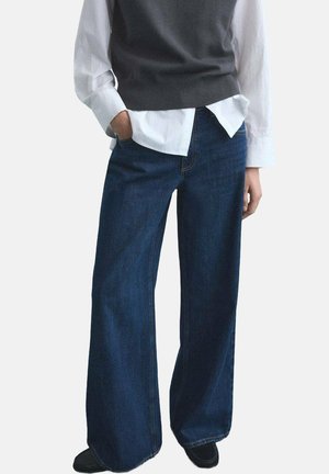 Person wearing wide-leg dark blue jeans, black shoes, white shirt, and dark gray sleeveless sweater with one hand in pocket.