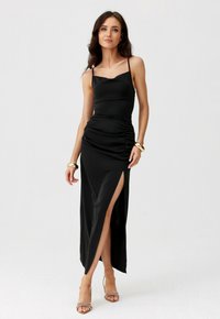 Žena v černých bezrukávových šatech s bočním rozparkem, zlatými náramky a béžovými sandály na vysokém podpatku, stojící před jednobarevným bílým pozadím.