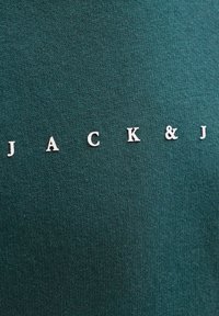 Lettere bianche che compongono "JACK & J" su uno sfondo di tessuto verde scuro e testurizzato.