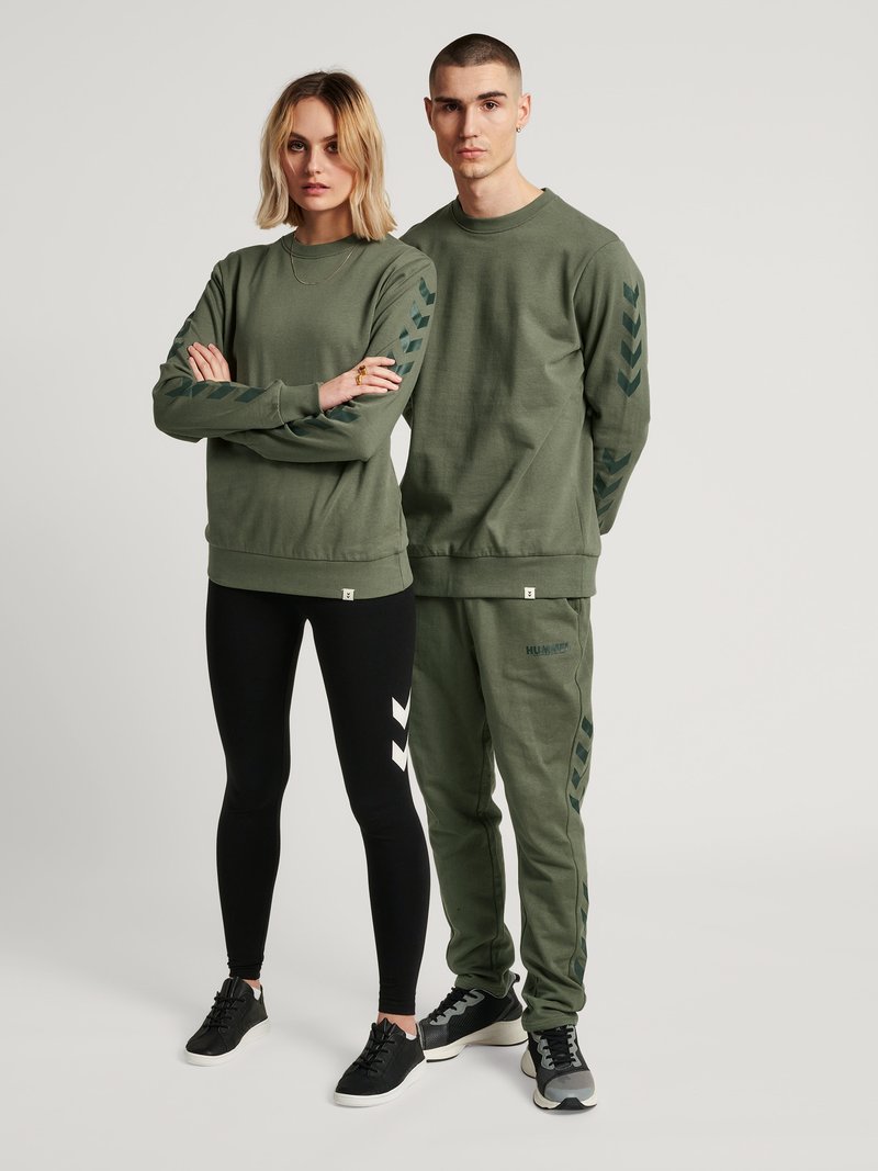 Twee volwassenen dragen bijpassende olijfgroene sweatshirts met pijlenpatronen op de mouwen; vrouw in zwarte legging met witte pijlen, beiden in sneakers.