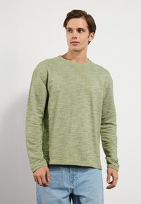 T-shirt à manches longues en vert clair, en tissu texturé. Présente une coupe décontractée et un design minimaliste. Associé à un jean en denim bleu clair.