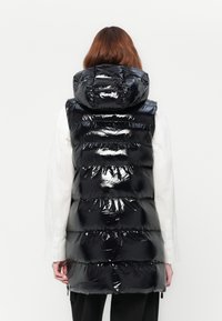Glanzend zwart puffer vest met capuchon, gedragen over een wit langemouwenshirt, getoond van achteren op een persoon met bruin haar.