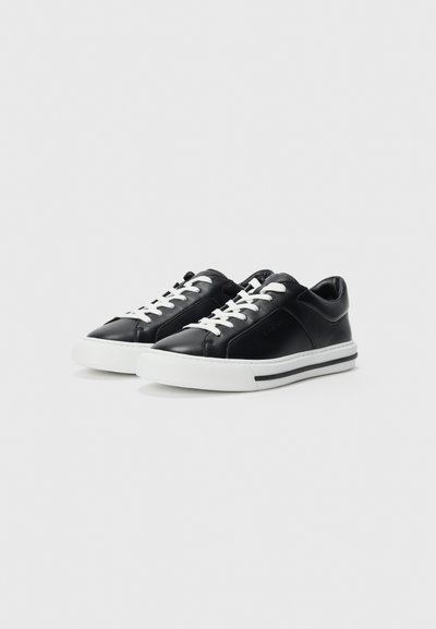Sneakers negre cu talpă joasă, cu șireți albi și tălpi groase albe, având o dungă neagră, expuse pe un fundal simplu, deschis la culoare.