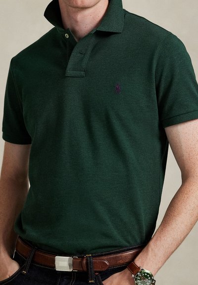 Homme portant un polo vert foncé avec un emblème violet, une ceinture en cuir marron, un jean foncé et une montre verte avec un bracelet marron.
