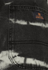 Tejido de mezclilla negra con patrones de tie-dye en gris claro. Presenta un bolsillo cosido y una etiqueta pequeña azul con texto en naranja que indica detalles de la marca y el estilo.