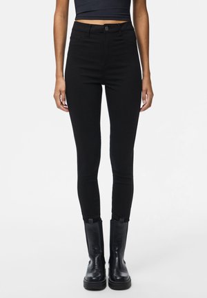 Vrouw die zwarte skinny jeans met hoge taille, een zwart cropped topje en zwarte leren laarzen tot halfkuit draagt, staand tegen een witte achtergrond.
