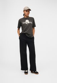 Νεαρός που φοράει γκρι μπλουζάκι με το λογότυπο «DREAMERS» και ένα λευκό λουλούδι, μαύρο φαρδύ παντελόνι, μαύρα αθλητικά παπούτσια και μπεζ καπέλο, στέκεται με τα χέρια στις τσέπες.