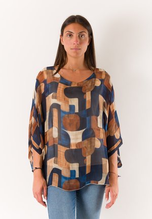 Donna con capelli lunghi e scuri che indossa una camicetta larga e colorata con motivo geometrico in tonalità di blu, marrone e beige, e jeans azzurri su sfondo bianco.