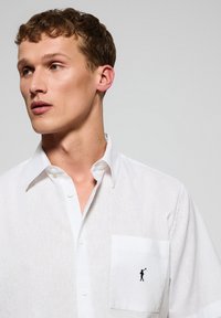 Camicia bianca in lino con colletto button-down, dotata di una tasca sul petto sinistro ricamata con una piccola figura nera che tiene in mano una tromba.