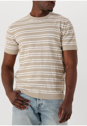 Man die een beige-wit horizontaal gestreepte trui met korte mouwen draagt en een lichtblauwe spijkerbroek, staat met één hand in de zak en kijkt naar rechts.