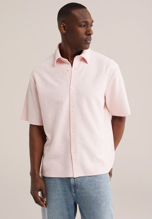 Homme portant une chemise à manches courtes rose clair boutonnée et un jean bleu, regardant vers sa droite sur un fond uni.