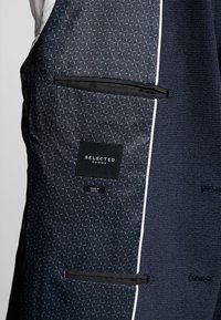 Inre foder av en marinblå mönstrad kavaj med två fickor och logotypen "SELECTED HOMME" sydd över "Made in China"-etiketten.