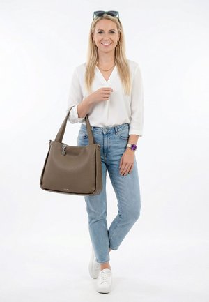 Mujer sonriente con blusa blanca y jeans azul claro sostiene un bolso de cuero marrón sobre el hombro, lleva zapatillas blancas y reloj púrpura.
