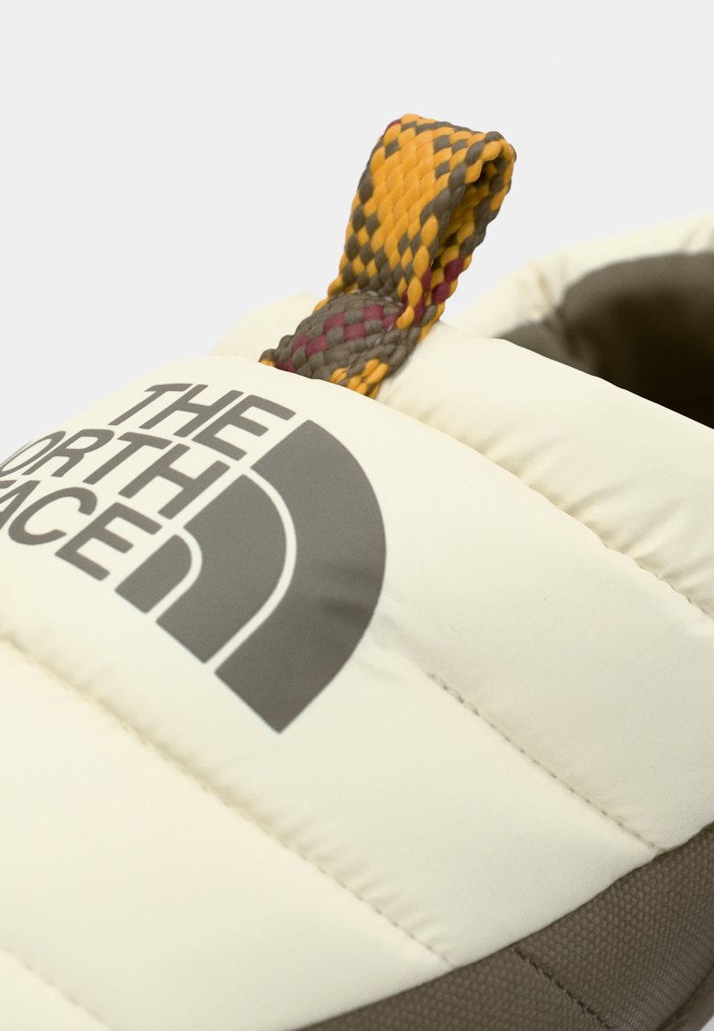 Gepolsterter Slipperschuh mit beigem Obermaterial und einem auffälligen grauen Logo, einer strukturierten braunen Sohle und einer mehrfarbigen gewebten Zugschlaufe.