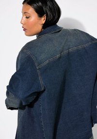 Chaqueta de denim en azul oscuro con un área de hombro deslavada, que presenta un cuello clásico, hombros caídos y detalles de costura visibles.