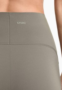 Leggings taille haute en tissu vert olive atténué. Les caractéristiques comprennent une texture lisse, des coutures marquées et un logo "OYSHO" sur la taille.