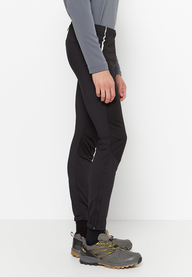 Pantaloni sportivi neri dal design aderente, con zip laterali e una texture liscia. Abbinati a scarpe da trekking grigie con dettagli gialli.