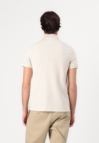 Beige kortærmet polo skjorte set fra ryggen, med klassisk krave, glat tekstur og sideslidser. Kombination med lyse bukser.