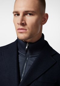 Granatowy płaszcz wełniany z mieszanki włókien z prążkowanym wysokim kołnierzem, z suwakiem z przodu i pikowaną podszewką. Gładka faktura i dopasowany krój.