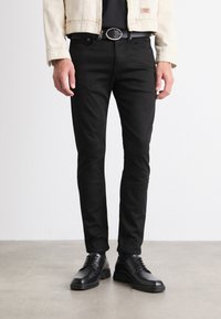 Jeans ajustados negros con una textura suave, que cuentan con cinco bolsillos y pasadores para cinturón, combinados con zapatos negros de cordones.