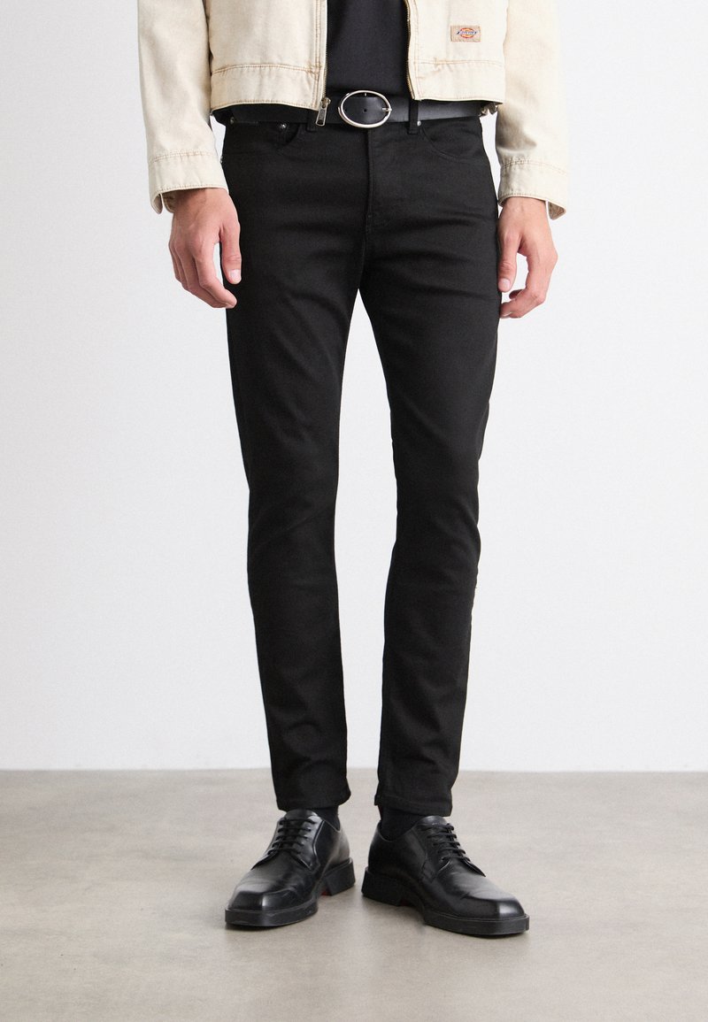 Jeans ajustados negros con una textura suave, que cuentan con cinco bolsillos y pasadores para cinturón, combinados con zapatos negros de cordones.
