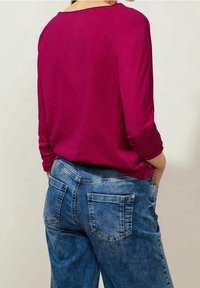 Fuchsia långärmad topp med rund halsringning, i kombination med blå denimjeans som har bakfickor och en något lös passform.