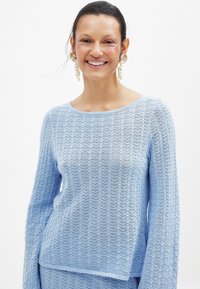 Nebesno modra, dolga rokava pletena majica z teksturo in širokim okroglim izrezom. Tkanina je lahka in nekoliko prosojna.