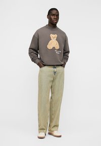 Άνδρας που στέκεται με τα χέρια στις τσέπες, φορώντας γκρι φούτερ με σχέδιο αρκουδάκι, μπεζ παντελόνι φαρδιάς γραμμής και λευκά παπούτσια σε λευκό φόντο.