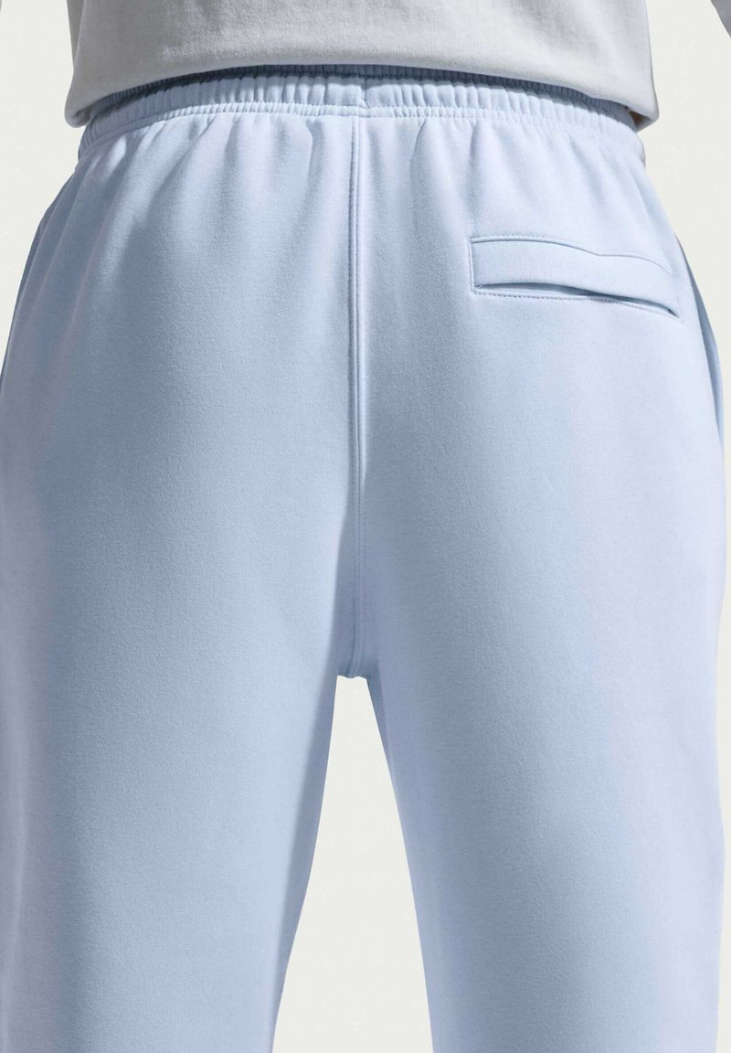 Persona che indossa pantaloni della tuta azzurri con fascia elastica in vita e una sola tasca posteriore, vista dalla vita in giù, retro.
