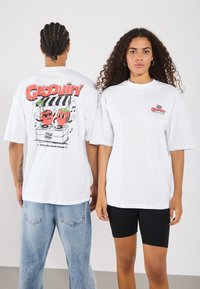 Hvid oversized t-shirt med et farverigt grafisk print af to animerede frugter på ryggen og et mindre logo på forsiden.