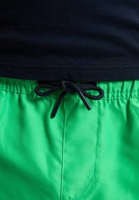 Pantaloni de baie verzi cu închidere prin șnur, având o textură netedă și un cordon negru contrastant. Tricou negru parțial vizibil deasupra.