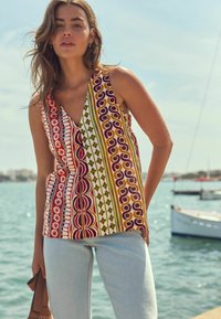 Top sans manches avec un décolleté en V, des motifs géométriques multicolores et une coupe décontractée, associé à un jean bleu clair et un sac à main.