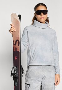 Svetlo modra, teksturirana, pulover z visokim ovratnikom iz flisa z žepom; sivi vzorčasti smučarski hlače. Model drži rjave smuči z vezmi.