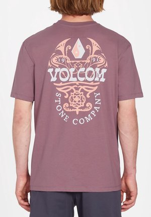 Personne portant un t-shirt mauve avec un grand logo beige et blanc de la Volcom Stone Company et une illustration décorative dans le dos.