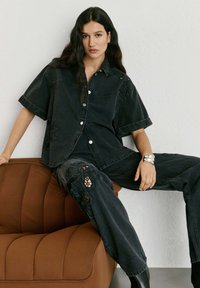 Frau mit langen dunklen Haaren, die auf einem braunen Stuhl sitzt, trägt ein schwarzes übergroßes Jeanshemd und Hosen mit Spitzenausschnitten, silbernes Armband am Handgelenk.