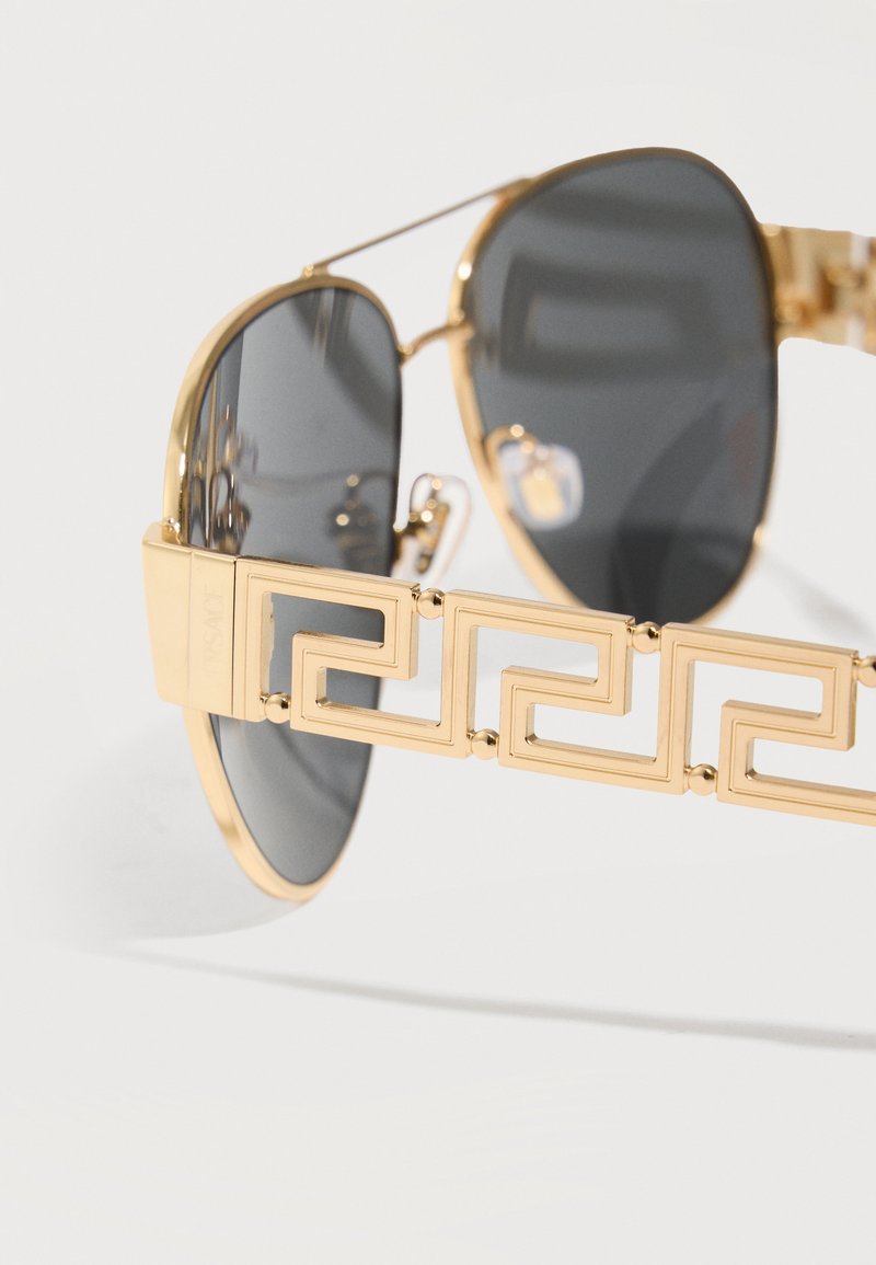 Lunettes de soleil aviateur avec montures dorées et verres foncés, présentant un motif géométrique doré détaillé sur les branches.